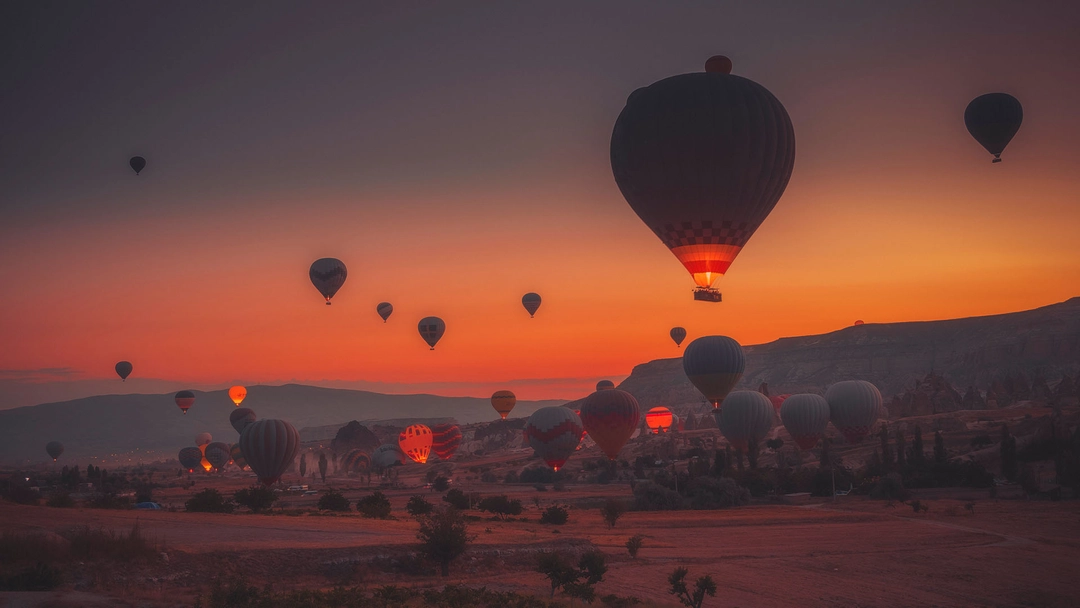 Numerous hot air balloons glowing and floating over a rocky landscape at sunset.